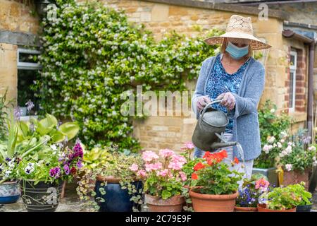 Frau, die Pflanzen und Blumen im Garten wässern und eine Gesichtsmaske während der Coronavirus Covid-19 Pandemie im Lockdown trägt Stockfoto