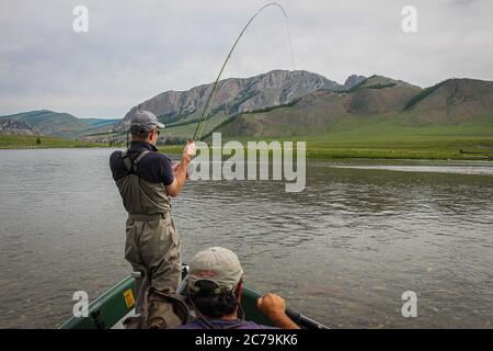 Ein Fliegenfischer mit einer Taimenforelle am Ende seiner Linie von einem Boot aus, auf dem Delger Moron Fluss in der Mongolei, Moron, Mongolei - 14. Juli 2014 Stockfoto
