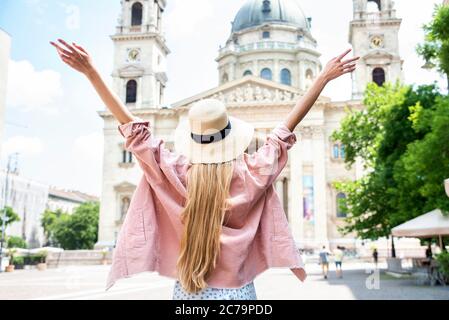 Rückansicht einer langhaarigen Frau mit Strohhut, während sie mit ausgestreckten Armen vor der St.-Stephans-Basilika in Budapest stand. Stockfoto