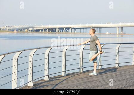 Sportlich reifer Mann hält Geländer und Stretching Bein am Flussufer während der Vorbereitung auf das Laufen Stockfoto