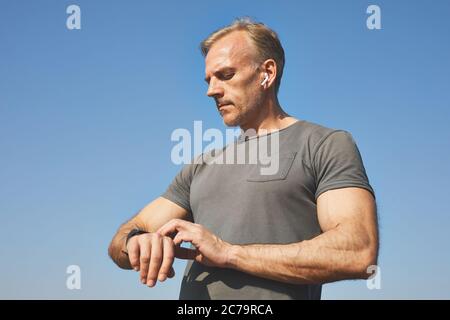 Selbstbewusster, schöner, reifer Mann in Ohrstöpseln, der gegen blauen Himmel steht und mit der Smartwatch den Puls der Zeit nimmt Stockfoto