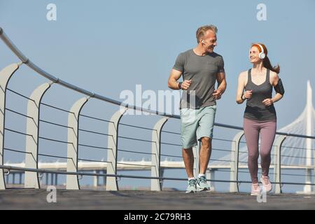 Positive aktive ältere Paar in Kopfhörer, die einander beim Joggen zusammen auf dem Damm zu sehen Stockfoto