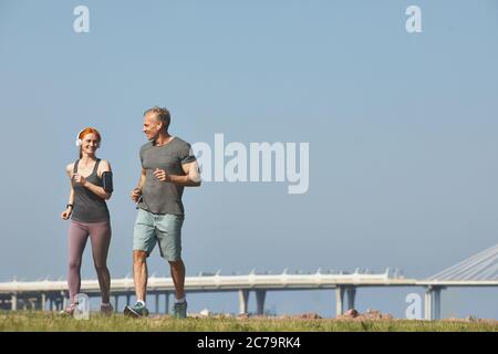 Sportliche Rothaarige Frau in kabellosen Kopfhörern joggen mit Freund und plaudern mit ihm Stockfoto