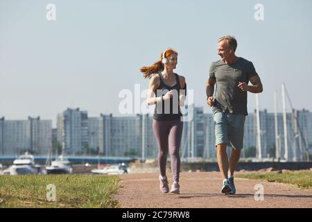Sportlich reifes Paar in Kopfhörern lächelt sich an, während sie gemeinsam in der Stadt joggen Stockfoto