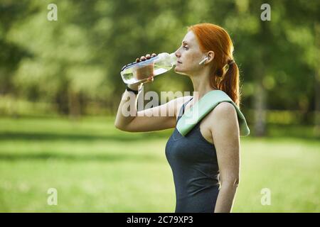 Seitenansicht der reifen Rothaarige Fitness-Frau mit Handtuch auf der Schulter Trinkwasser im Park nach dem Training Stockfoto