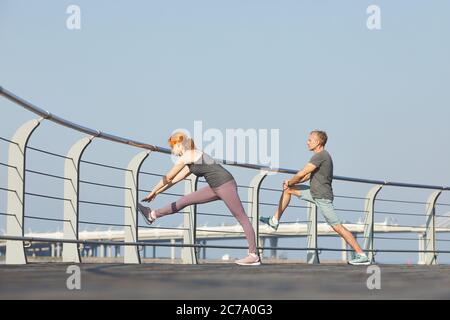 Sportlich modernes reifes Paar mit Geländer am Damm beim Dehnen der Beinmuskulatur vor dem Laufen Stockfoto