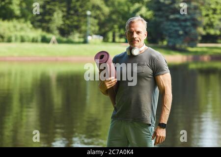 Portrait of content muskulöser reifer Mann mit Kopfhörern um den Hals hält Übungsmatte gegen Park See Stockfoto