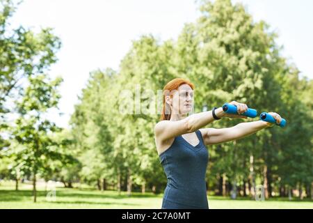 Fokussierte reife Rothaarige Frau mit Hanteln für Arm Übungen im Sommer Park Stockfoto