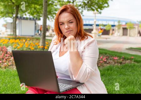 Freiberuflich. Kaukasische nachdenkliche Frau, die an einem Laptop in einem Park arbeitet. Plus-Größe. Konzept von Remote-Arbeit und Online-Geschäft. Stockfoto
