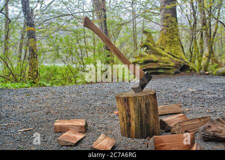 Eine Axt eingebettet in einen Holzstumpf mit frisch gehacktem Brennholz, das sie auf einem kiesbedeckten Campingplatz umgibt. Stockfoto