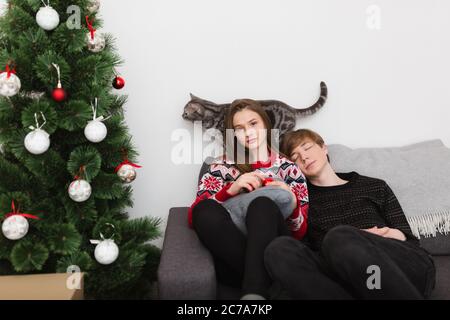 Portrait von schönen Mädchen auf dem Sofa sitzen und traumhaft in der Kamera suchen, während Junge auf sie lehnt und schlafen mit Katze und Weihnachtsbaum auf Stockfoto
