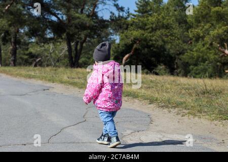 Kleines einsames Kind in einer leuchtend rosa Jacke geht auf einer asphaltierten Straße im Freien Stockfoto