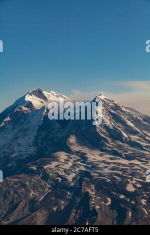 Schneebedeckter vulkanischer Berg mit einer Rauchwolke, die über den Himmel strömt und die dramatische und zerklüftete Wildnis Alaskas zeigt. Stockfoto