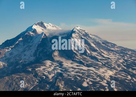 Schneebedeckter vulkanischer Berg mit einer Rauchwolke, die über den Himmel strömt und die dramatische und zerklüftete Wildnis Alaskas zeigt. Stockfoto