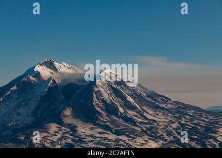 Schneebedeckter vulkanischer Berg mit einer Rauchwolke, die über den Himmel strömt und die dramatische und zerklüftete Wildnis Alaskas zeigt. Stockfoto