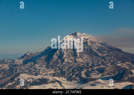 Schneebedeckter vulkanischer Berg mit einer Rauchwolke, die über den Himmel strömt und die dramatische und zerklüftete Wildnis Alaskas zeigt. Stockfoto