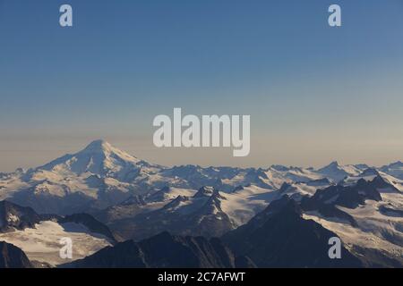 Schneebedeckter vulkanischer Berg mit einer Rauchwolke, die über den Himmel strömt und die dramatische und zerklüftete Wildnis Alaskas zeigt. Stockfoto