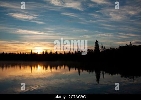 Goldener Sonnenuntergang über einem See Alaskas, mit Bergsilhouetten und weichen Wolken, die die ruhige Schönheit der Wildnis während der Dämmerung widerspiegeln. Stockfoto