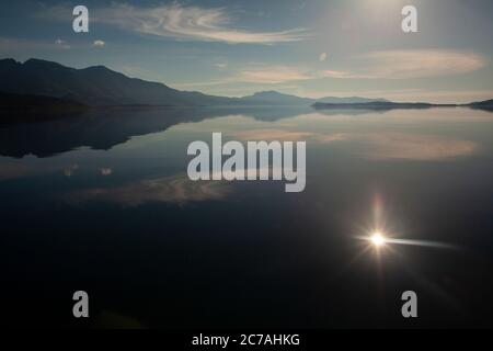 Der Iliamna-See mit spiegelähnlichen Reflexen des Himmels und der Berge, während die Sonne ein strahlendes Licht über das unberührte Wasser der Wildnis wirft. Stockfoto
