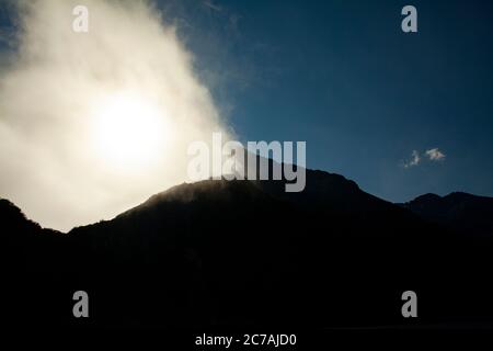 Nebel rollt über das Wasser des Lake Iliamna, Alaska, und die Sonne strahlt Licht über die zerklüftete Bergsilhouette und die ruhige Wildnis Landschaft Stockfoto