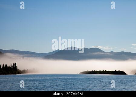 Der Morgennebel zieht über den Lake Iliamna, Alaska, mit Bergen im Hintergrund und Sonnenlicht, das sich vom ruhigen, ruhigen Wasser der Wildnis reflektiert Stockfoto