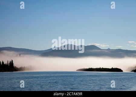 Der Morgennebel zieht über den Lake Iliamna, Alaska, mit Bergen im Hintergrund und Sonnenlicht, das sich vom ruhigen, ruhigen Wasser der Wildnis reflektiert Stockfoto