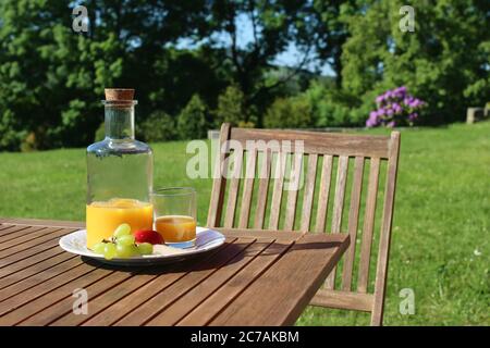 Flasche und Glas Orangensaft auf Holztisch mit Stuhl. Verschwommene grüne Bäume und Hügel Gipfel Hintergrund. Frühstück im Freien mit Obst auf dem Land Stockfoto