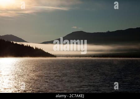 Der Morgennebel zieht über den Lake Iliamna, Alaska, mit Bergen im Hintergrund und Sonnenlicht, das sich vom ruhigen, ruhigen Wasser der Wildnis reflektiert Stockfoto