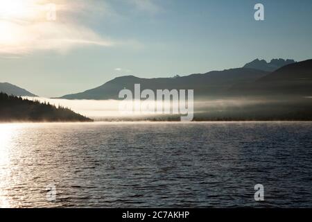 Der Morgennebel zieht über den Lake Iliamna, Alaska, mit Bergen im Hintergrund und Sonnenlicht, das sich vom ruhigen, ruhigen Wasser der Wildnis reflektiert Stockfoto