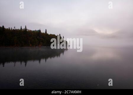 Ein nebeliger Morgen über dem Lake Iliamna, Alaska, mit ruhigem Wasser, das die bewaldete Küste und die nebelige Atmosphäre reflektiert und die ruhige Wildnis eingefangen Stockfoto