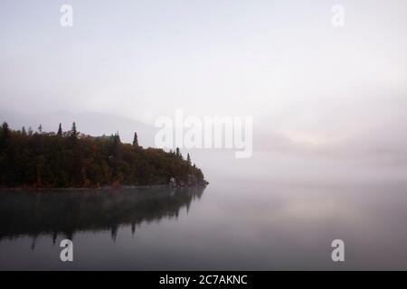 Ein nebeliger Morgen über dem Lake Iliamna, Alaska, mit ruhigem Wasser, das die bewaldete Küste und die nebelige Atmosphäre reflektiert und die ruhige Wildnis eingefangen Stockfoto