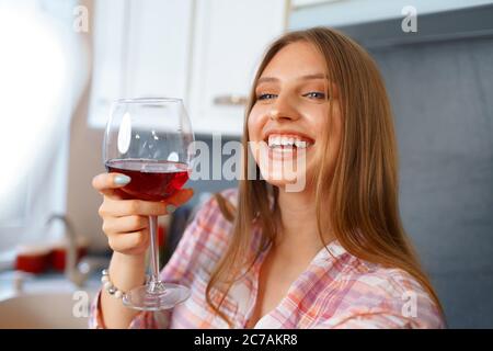 Glückliche, entspannte junge Frau, die mit einem Glas Rotwein in der Küche steht und ihr Smartphone benutzt Stockfoto