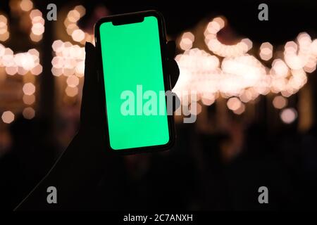 Nahaufnahme mit einer Hand und Smartphone mit grünem Bildschirm in der Nacht. Schöne goldene Bokeh Hintergrund Stockfoto
