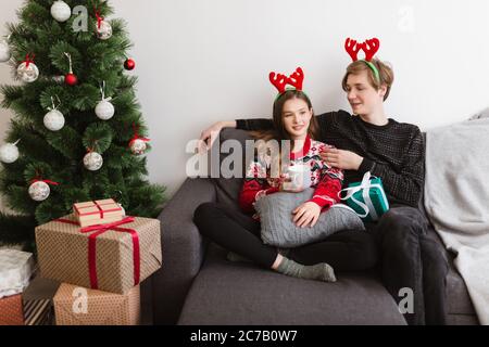 Junge schöne Paar sitzt auf dem Sofa zu Hause und trägt Hirsche Hörner, während die Zeit zusammen mit Weihnachtsbaum in der Nähe Stockfoto
