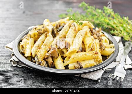 Penne Pasta mit Pilzen in einem Teller auf einem Küchentuch, Thymian, Gabel und Knoblauch auf schwarzem Holzbrett Hintergrund Stockfoto