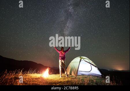 Weibliche Wanderer, die sich am Sommernachtcamping neben dem Lagerfeuer und dem glühenden Touristenzelt ausruhen. Rückansicht einer jungen Frau, die die Hände hochhält und den Blick auf den Nachthimmel voller Sterne und Milchstraße genießt. Stockfoto