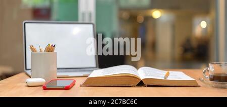Nahaufnahme des Lerntisches mit geöffnetem Buch, Laptop, Smartphone, Schreibwaren und Kaffeetasse auf Holztisch