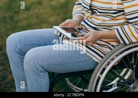 Modisch gekleidete Frau im Rollstuhl mit einem modernen Tablet in der Hand geht auf der Straße. Unkenntlich Person. Stockfoto
