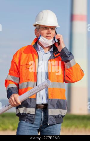 Ingenieur oder Architekt, der eine Gesichtsmaske aus Coronavirus und Blaupause in der Hand auf einer Baustelle trägt, die am Handy spricht Stockfoto