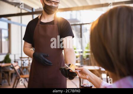 Der Kellner arbeitet in einem Restaurant in einer medizinischen Maske, Handschuhe während Coronavirus Pandemie. Stellt eine neue Norm in Service und Sicherheit dar. Bekommt bezahlte Bestellung mit Gerät kontaktlos. Leerer Bildschirm für Ihre Anzeige. Stockfoto