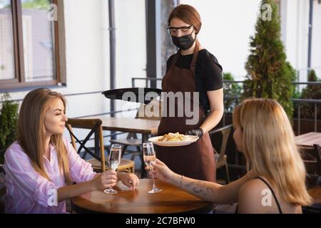 Die Kellnerin arbeitet in einem Restaurant in einer medizinischen Maske, Handschuhe während Coronavirus Pandemie. Stellt eine neue Norm in Service und Sicherheit dar. Bestellung aufgeben, Betreuung von Kunden, Besuchern. Stockfoto