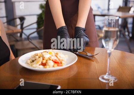 Die Kellnerin arbeitet in einem Restaurant in einer medizinischen Maske, Handschuhe während Coronavirus Pandemie. Stellt eine neue Norm in Service und Sicherheit dar. Bestellung, Bedienung, Betreuung von Kunden, Besuchern. Stockfoto