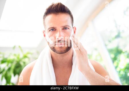 Nahaufnahme eines hübschen jungen Mannes mit einem Handtuch im Nacken, der vor dem Spiegel steht und Rasierschaum auf sein Gesicht aufgibt. Stockfoto