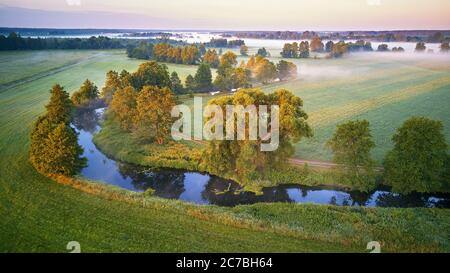 Sonnige Sommer ruhige Morgen Panorama. Kleiner Fluss, Nebel Sonnenaufgang Luftbild. Erste Sonnenstrahlen über grüne Wiese und Feld Stockfoto