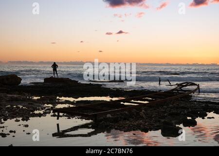 Silhouette einer Frau, die auf das Mittelmeer schaut, in der Nähe eines verlassenen Metallpiers am Strand, bei Sonnenuntergang, in Batroun, Libanon, im Mittleren Osten Stockfoto