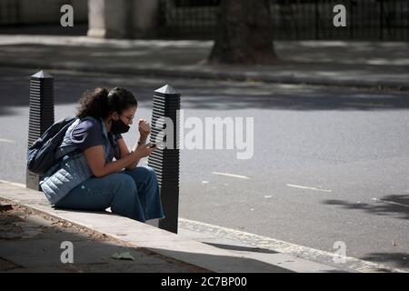 Eine junge Frau mit einem Pferdeschwanz, der eine Gesichtsmaske trägt, sitzt am Straßenrand und blickt auf ihr Telefon außerhalb der königlichen Gerichtshöfe. London, England, Großbritannien Stockfoto