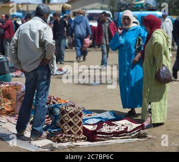 Marokkanische Frauen mit Djellaba auf der Suche nach geeigneten Kleidung, Stoffen und Teppichen auf einem Markt (Souk) in einem Dorf in der Nähe von Fes in Marokko Stockfoto