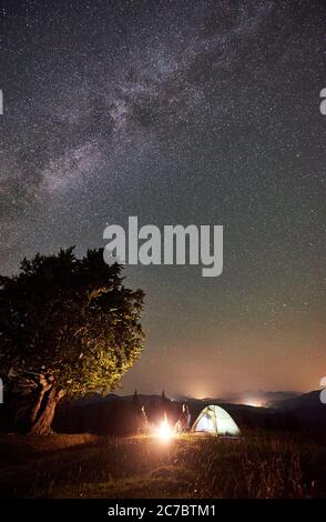 Junge Paar Touristen ruhen sich bei Sommernacht Camping in den Bergen. Mann und Frau sitzen auf Stühlen neben Lagerfeuer, beleuchtetes Zelt und großer Baum unter Sternenhimmel voller Sterne und Milchstraße. Stockfoto