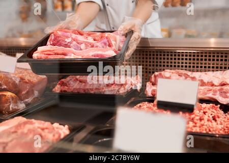 Nahaufnahme von rohen Fleischstücken im Kühlschrank mit Preisschildern bereit zum Verkauf in Fleisch Abteilung des Ladens. Weibliche Hände, die aus dem Glas Theke Teller mit Scheiben frisch Steaks. Konzept der Nahrung. Stockfoto