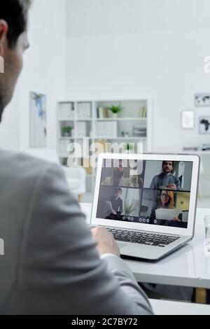 Über die Schulter schauen Sie, wie ein Geschäftsmann am Schreibtisch im Büro sitzt und mit Kollegen über Videokonferenzen auf einem Laptop spricht Stockfoto